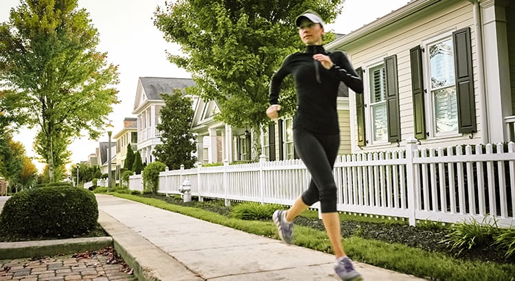 A person jogging on a sidewalk in a suburban neighborhood. The person is wearing athletic clothing and running shoes.