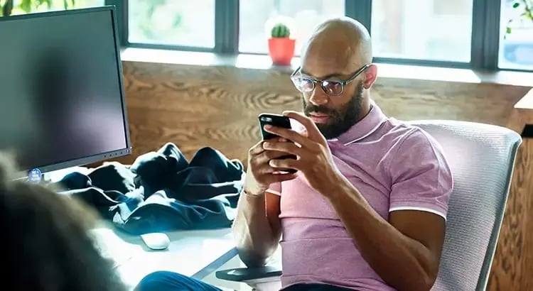 A man sitting and looking at his smartphone. He is wearing glasses and a casual outfit, with a serious expression on his face.