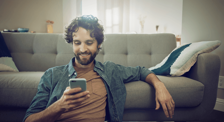 Man sitting on floor with back against a beige couch smiling and reading an article on this phone