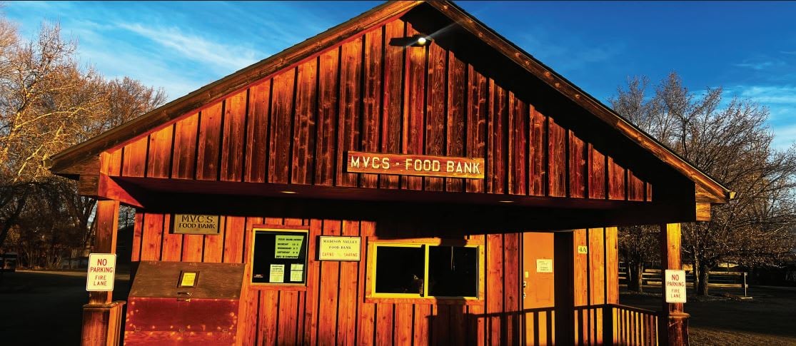 A close-up shot of a rustic wooden building with a sign that reads "MVCS FOOD BANK" and smaller text "MADISON VALLEY COMMUNITY SERVICE, INC."