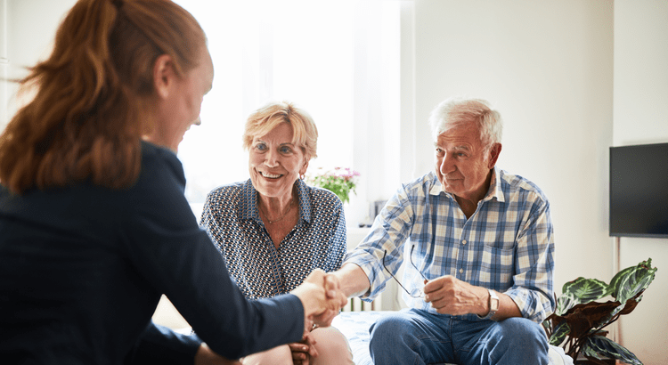 An informal meeting scene with a woman in business attire talking to an elderly couple. The woman appears to be giving advice or consultation, and the setting is a bright room with natural light.