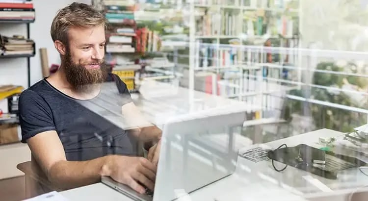 A man with a beard and glasses sitting at a desk, working on a computer. The setting appears to be a modern office or workspace, indicating productivity.