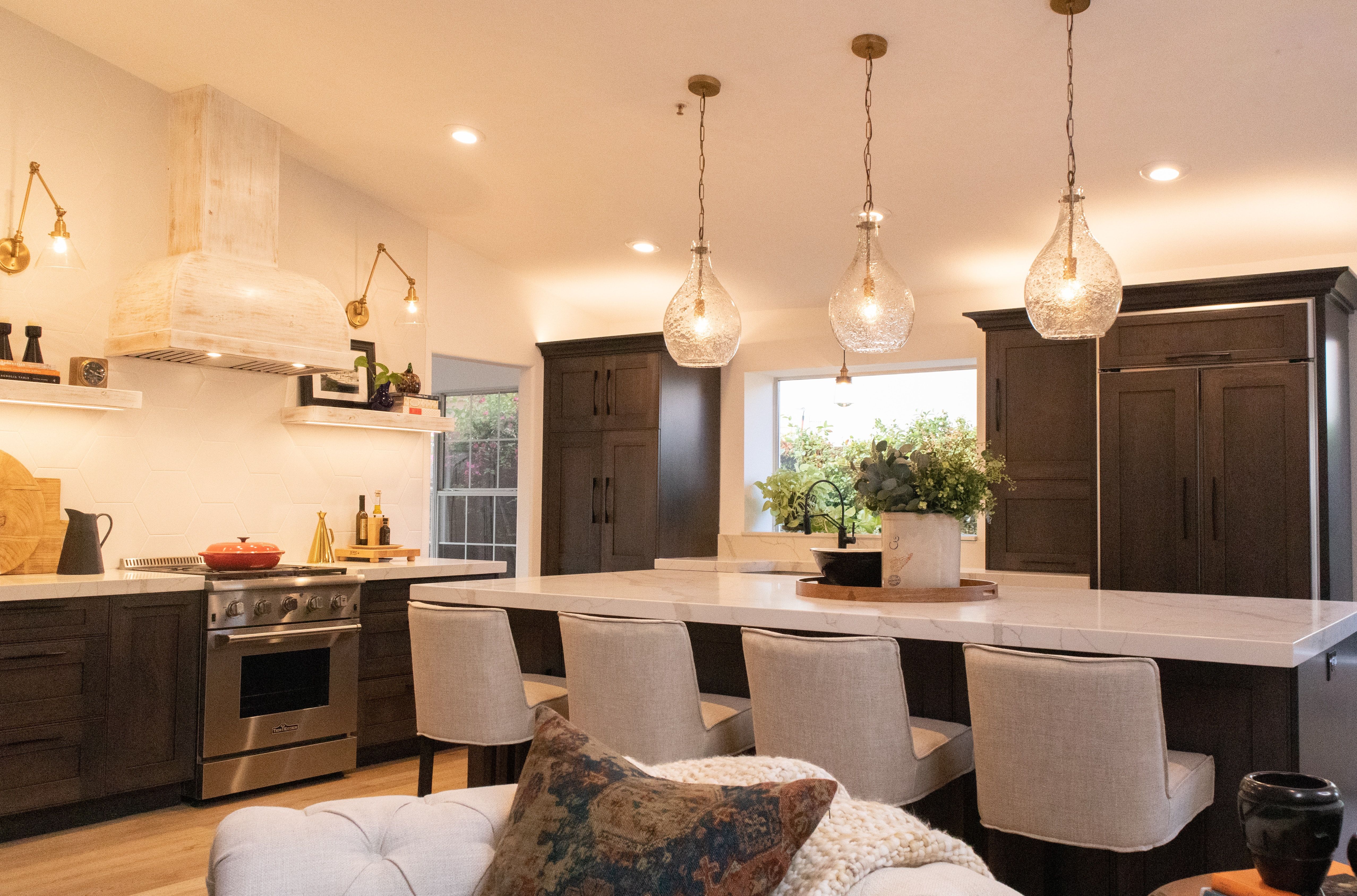Kitchen with dark cabinetry, marble island, upholstered chairs, and glass pendant lights.