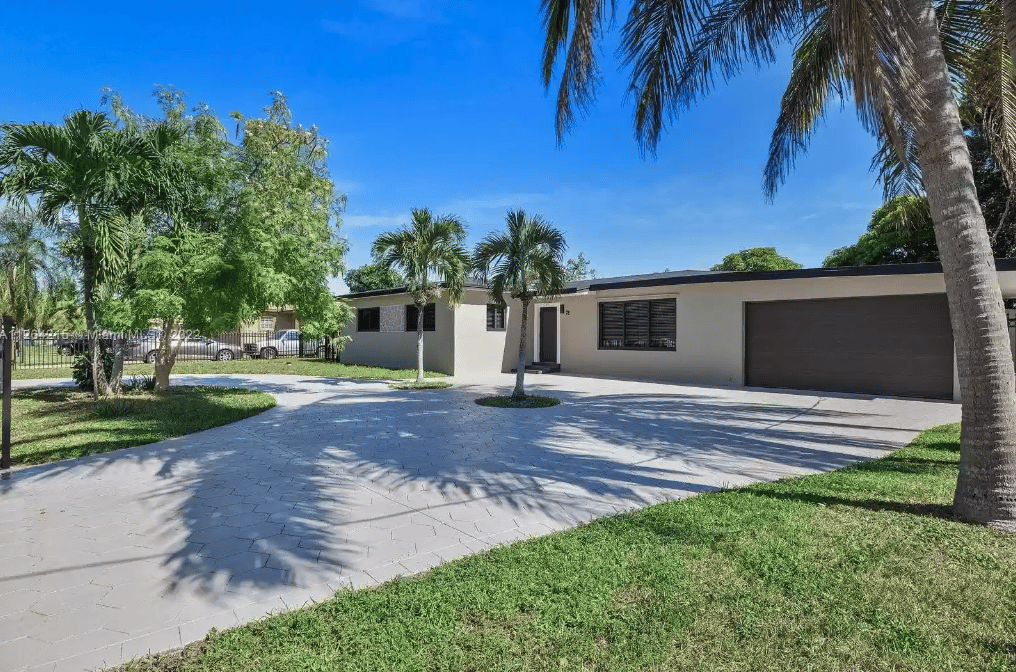 Single-story modern home with a broad driveway, dark garage door, and palm trees.