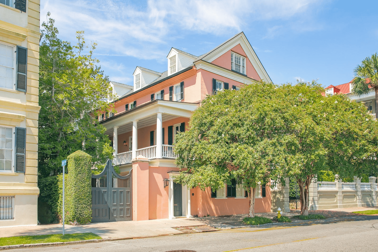 Pink Charleston home with white porches, dark shutters, and a gated entrance beneath mature trees.