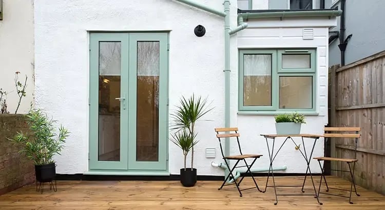 An exterior view of a house with a patio area. The patio features a small table, chairs, potted plants, and double glass doors leading inside.