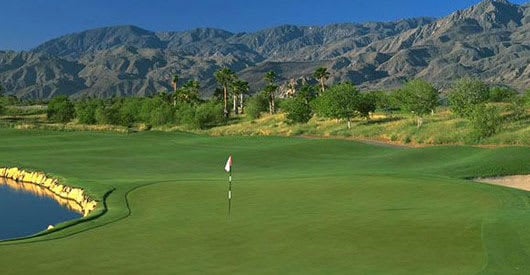 A golf course green with a flag and a pond in the foreground.