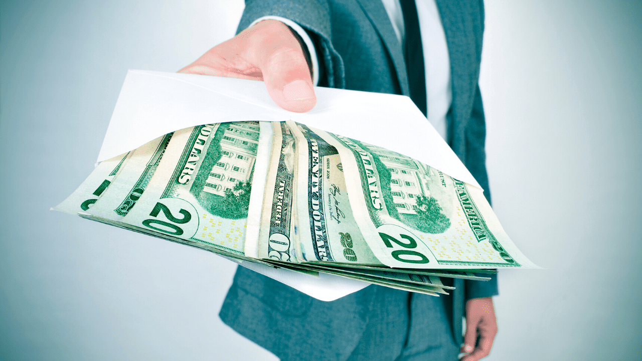 A person in a suit handing over an envelope filled with U.S. dollar bills. The envelope is open, revealing several twenty-dollar bills.