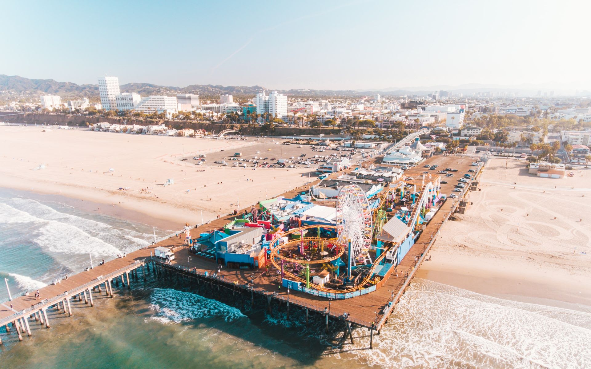 Aerial view of a beach pier with an amusement park, Ferris wheel, ocean waves, and sandy shoreline.