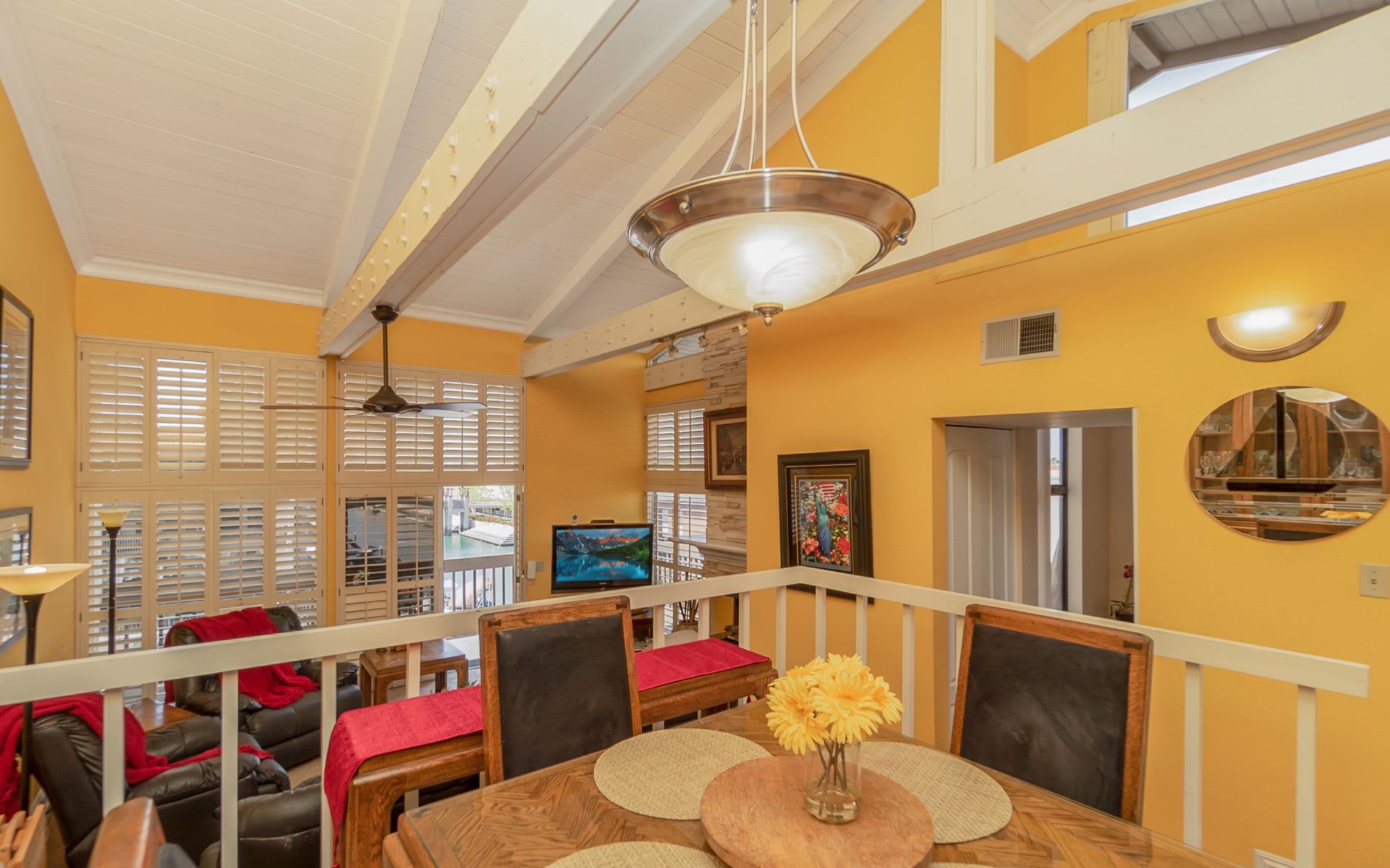 Furnished dining area overlooking a living room, with a vaulted beamed ceiling and plantation shutters.