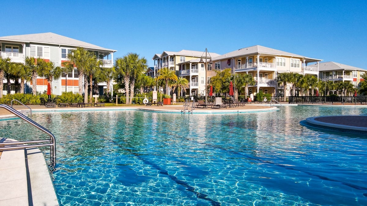 A large, rectangular swimming pool surrounded by colorful buildings and palm trees.