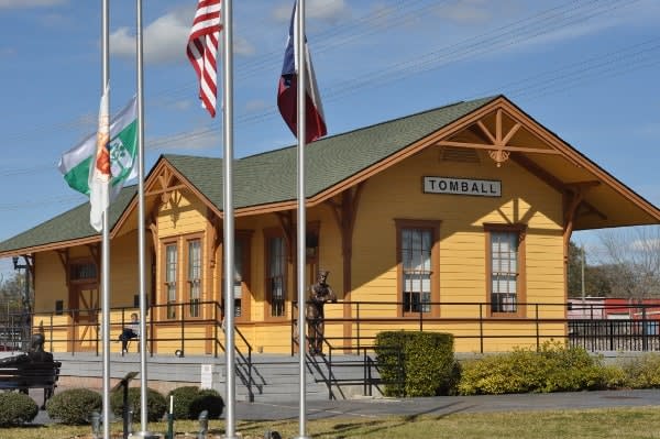 Yellow Tomball train depot with flags on three flagpoles and a gabled roof.