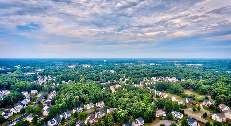 An aerial view of a suburban area with many houses, trees, and streets. The view extends to the horizon, showing a well-planned residential community.