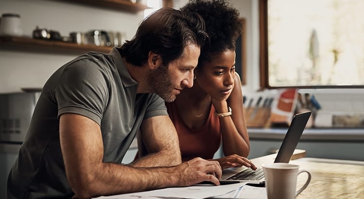 A man and a woman, possibly a couple, are sitting together, focused on a laptop. They appear to be engaged in a discussion, with coffee cups nearby, suggesting an intimate, thoughtful setting, possibly related to work or planning.