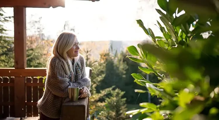 A woman standing on a porch, holding a cup and looking out at the garden. She is dressed warmly, indicating a cool day.