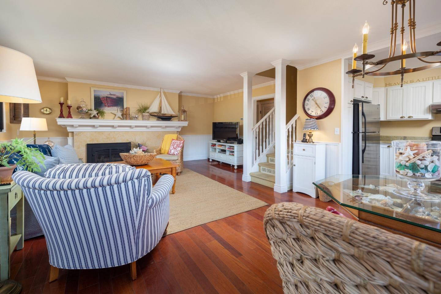 Living room with a stone fireplace, striped seating, hardwood floors, and an adjoining kitchen.