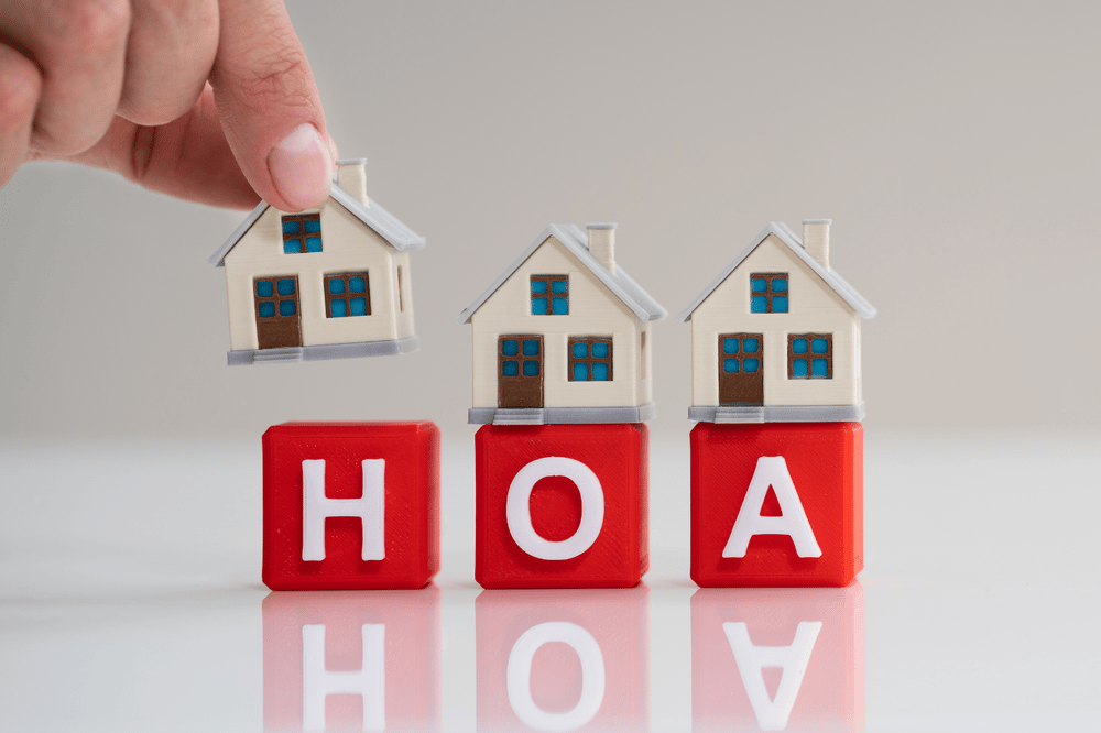 A hand places a miniature house beside two homes above red HOA letter blocks.