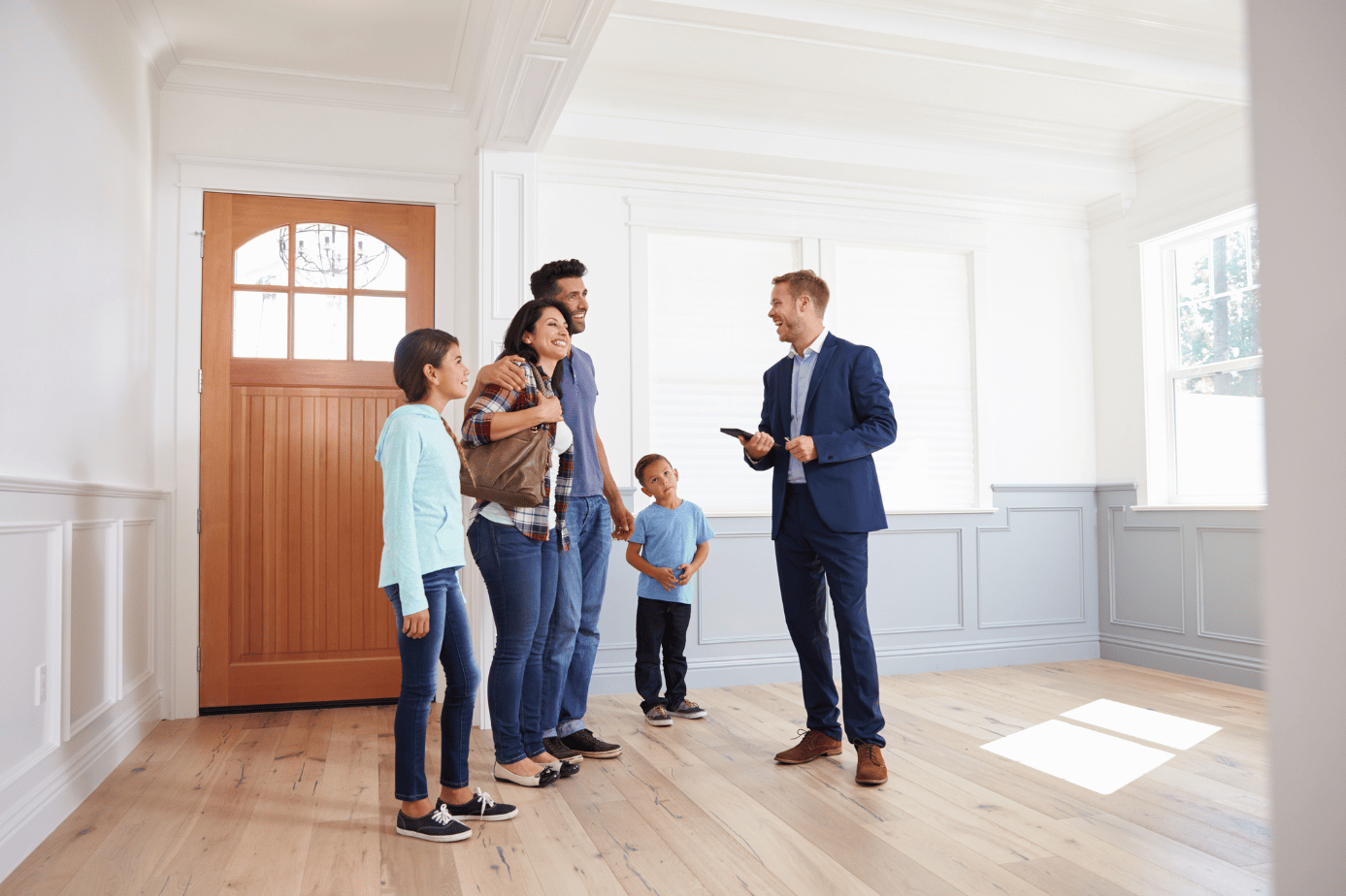 An agent shows a family a bright, unfurnished room with pale wood floors and a wooden entry door.