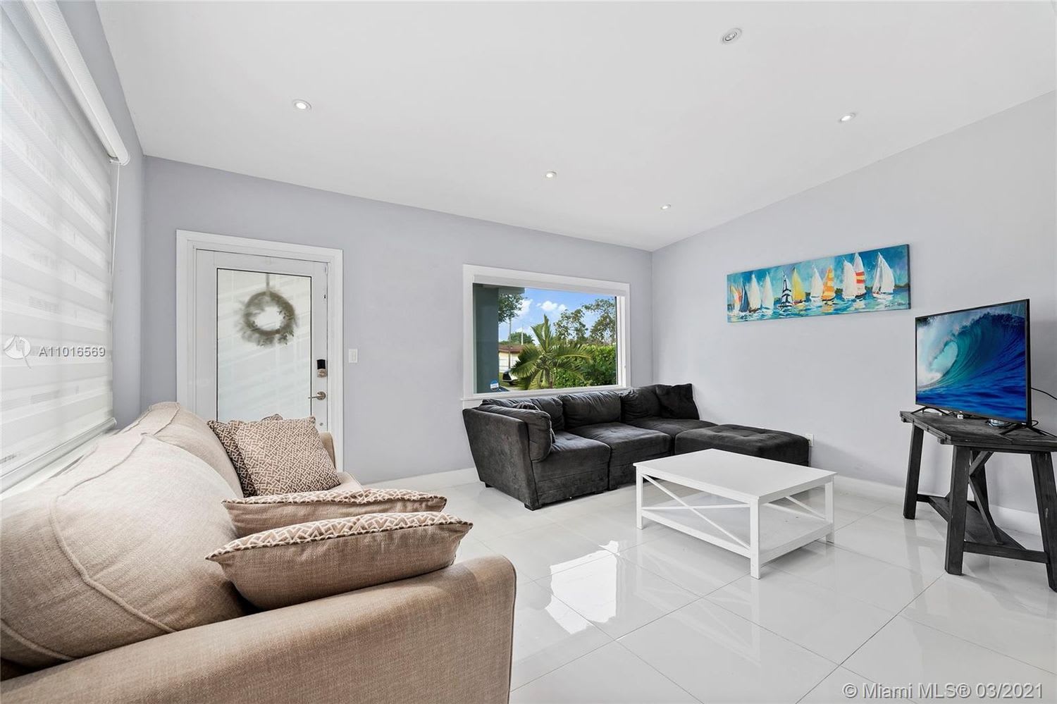 Living room with a dark sectional, beige sofa, glossy tile floors, and a window overlooking palm trees.