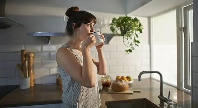 A woman standing in a kitchen, drinking a glass of water. The kitchen has a modern design with various kitchen appliances and utensils visible.