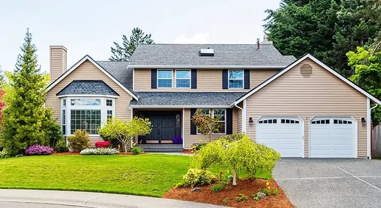 A suburban house with a well-manicured lawn, colorful flowers, and a driveway leading to a two-car garage. The house has a gabled roof and a mix of brick and siding exterior.