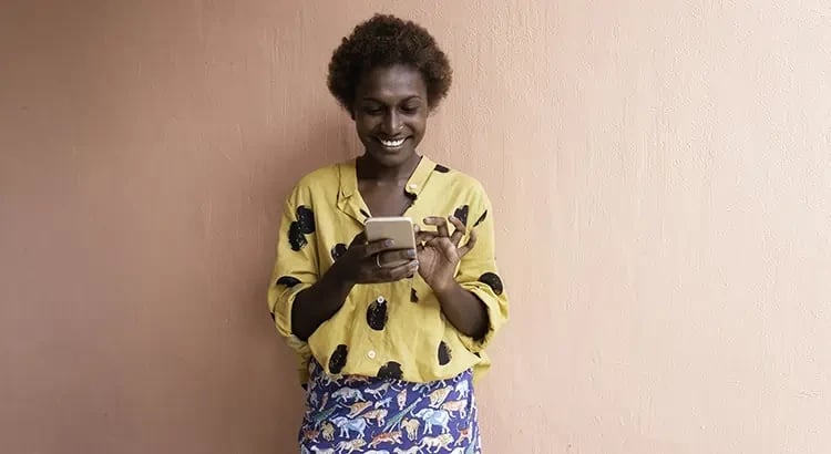 A woman in a yellow top and a floral skirt standing against a peach-colored wall, smiling while looking at her phone.