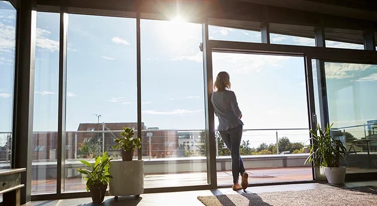 A woman standing by large windows in a modern building, possibly representing real estate opportunities, professional life, or decision-making.