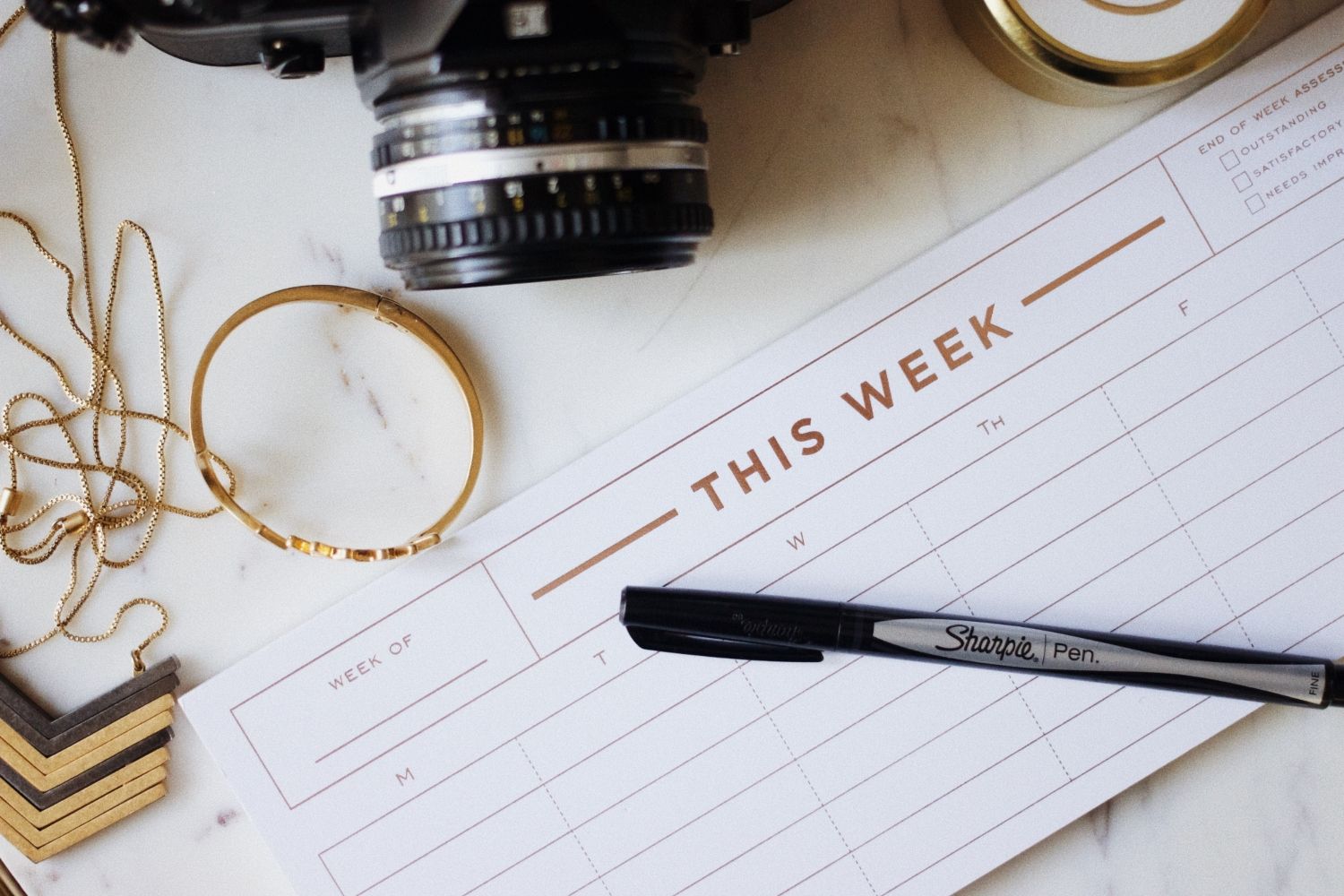 Weekly planner and Sharpie pen beside a camera lens, gold-framed glasses, a watch, and chain.