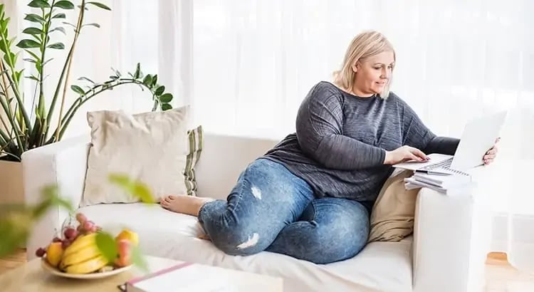 A woman sitting comfortably on a couch with a laptop on her lap. She is dressed in casual attire, indicating a relaxed, home-working environment.