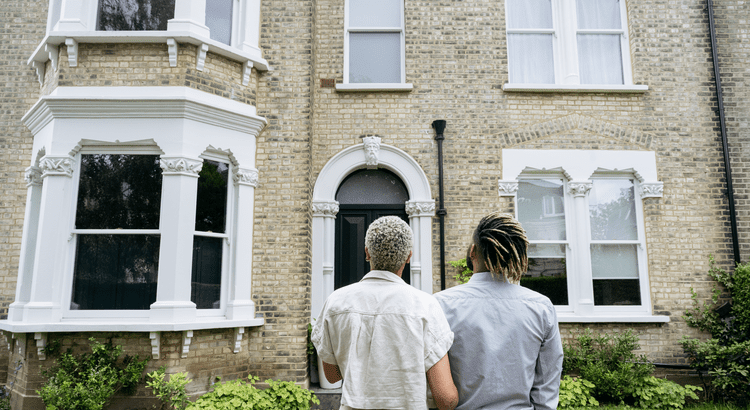 Two people standing close together, facing the front of a brick house with white trim and large windows. 