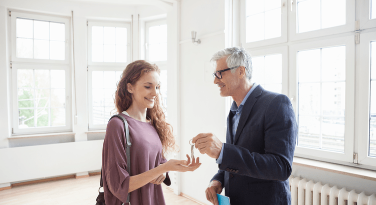 A young woman and an older man standing in a bright, modern room with large windows. The man, dressed in a suit, is handing a key to the woman, who is smiling and extending her hand to receive it. 