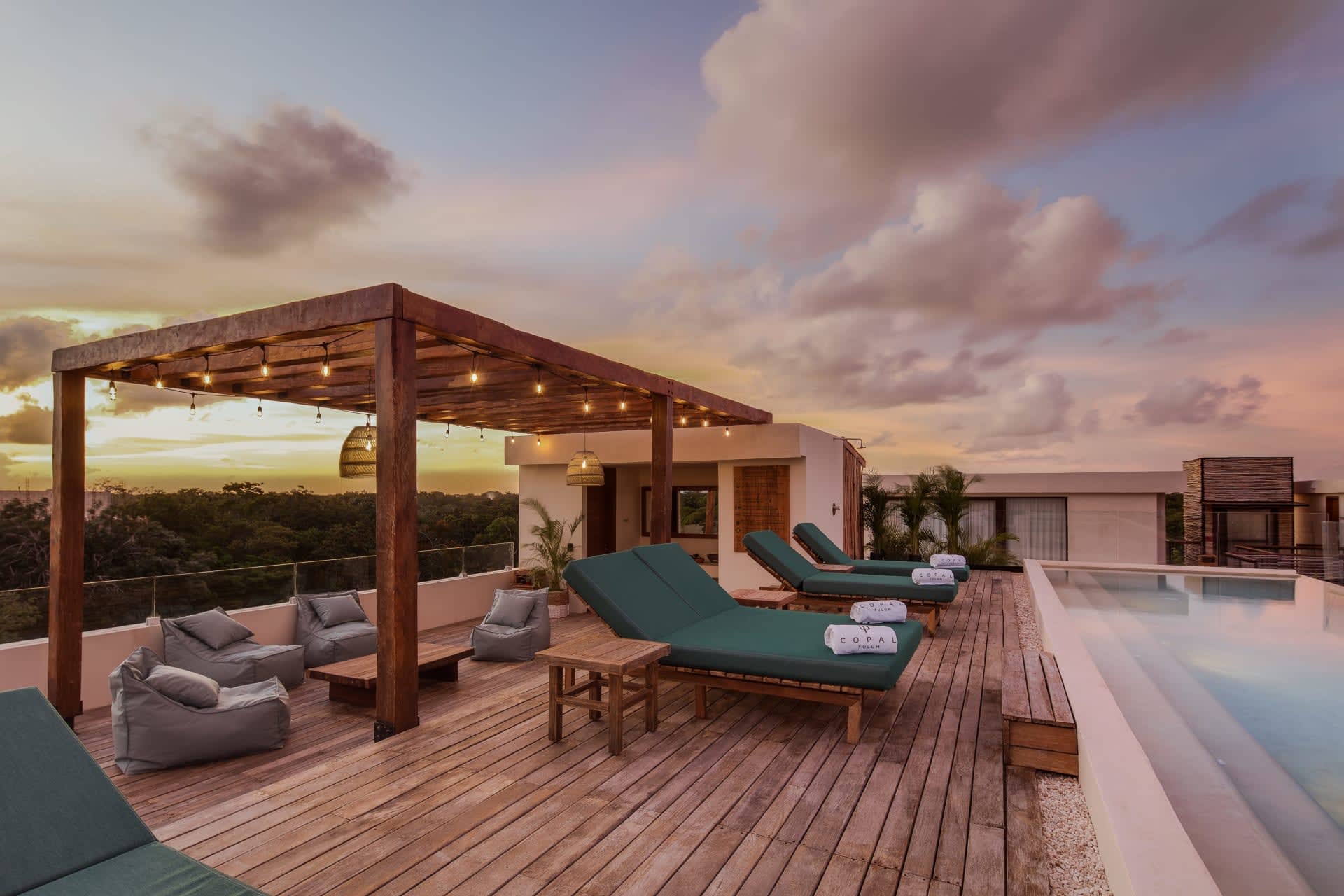 Rooftop pool terrace with a wooden pergola, lounge chairs, and sunset views over treetops.