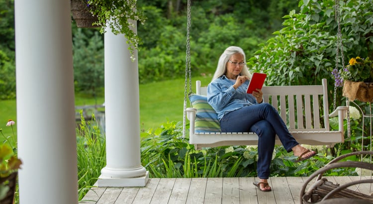 A senior woman sitting on a porch, reading a book.