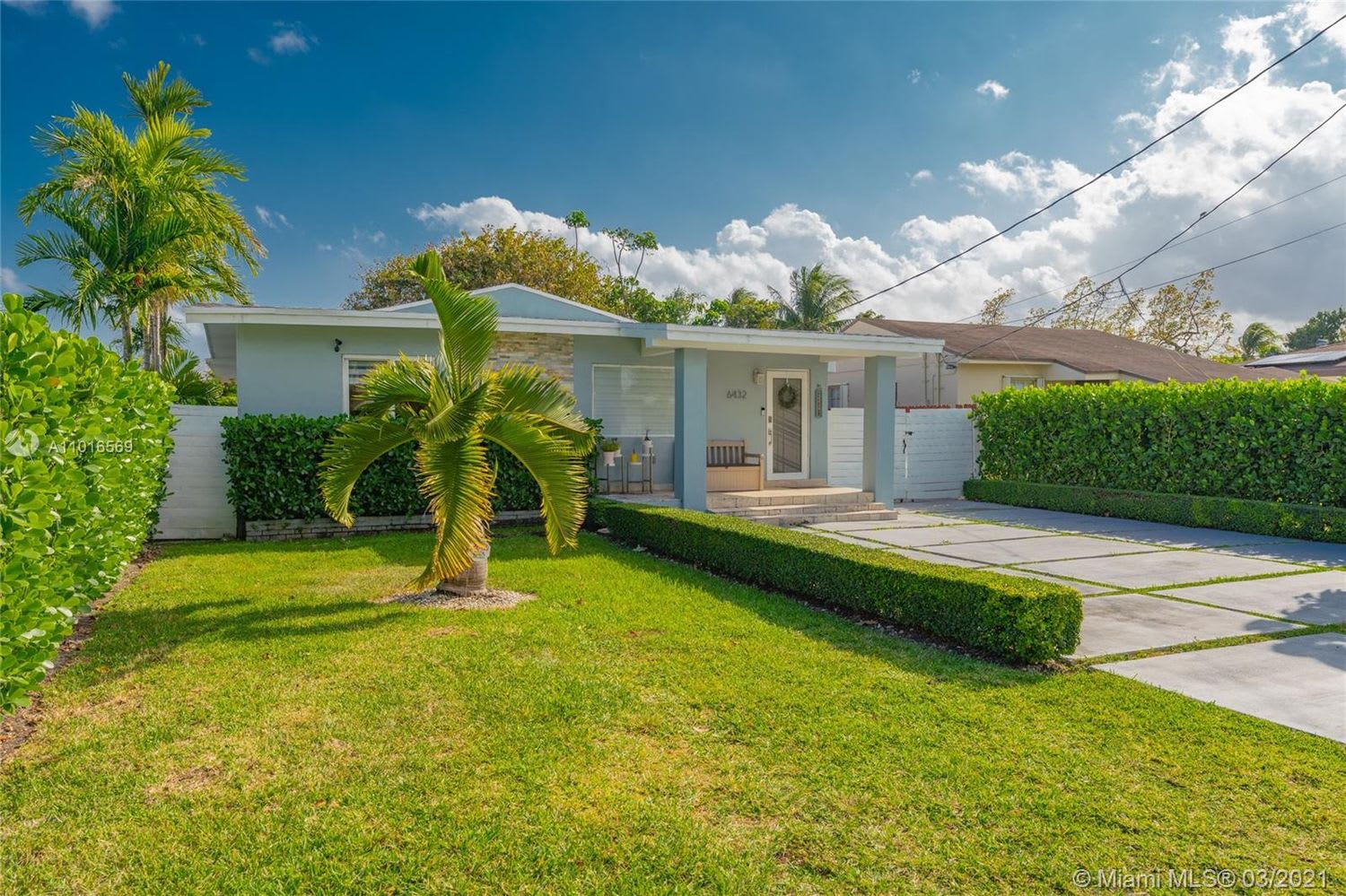 Light blue single-story house with a covered entry, palm trees, hedges, and a paved driveway.