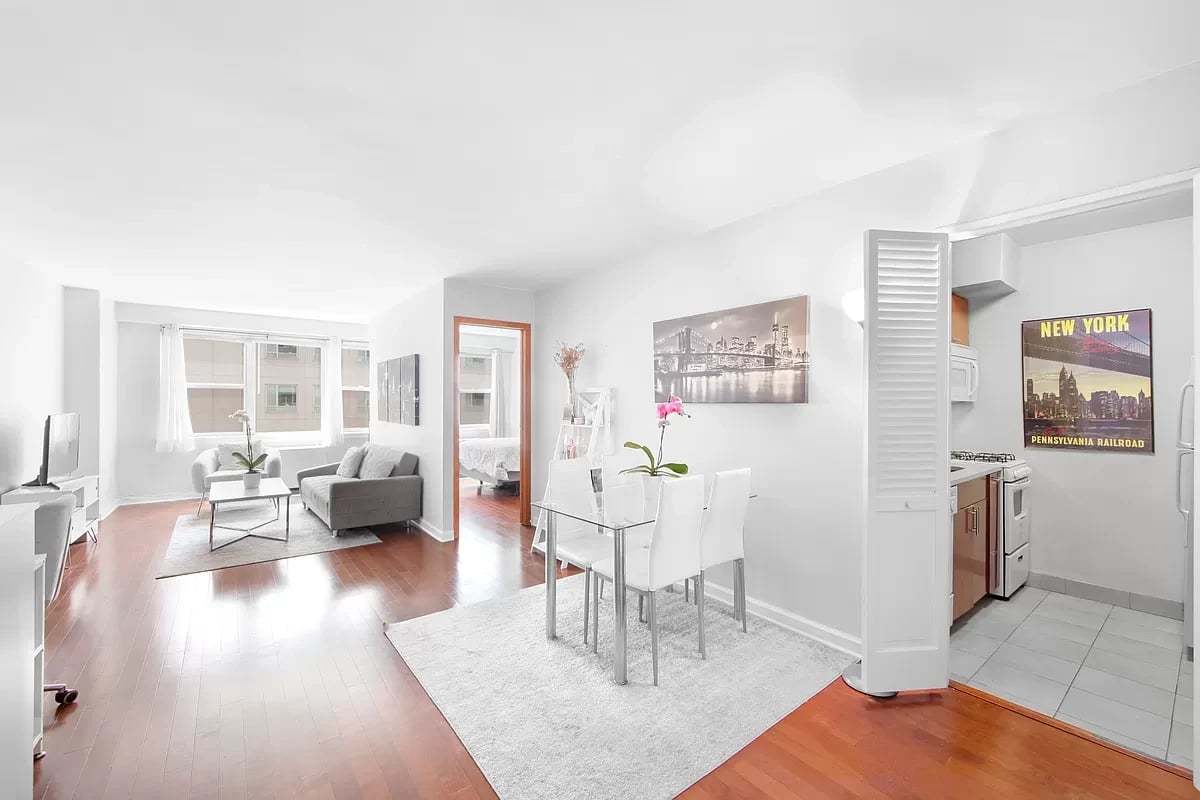 Bright open living and dining area with a gray sofa, glass table, hardwood floors, and adjoining kitchen.