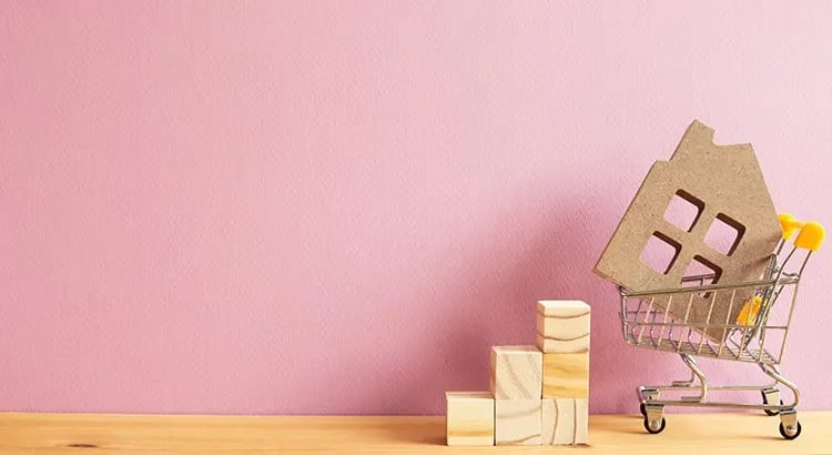 A small house cutout in a shopping cart, placed against a pink background. Next to the cart are wooden blocks, possibly representing home-related investments or purchases.