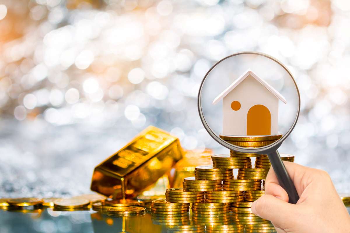 A magnifying glass highlights a small house atop stacks of gold coins.