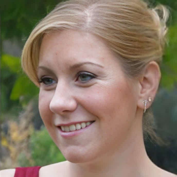 A woman with light blonde hair styled in an updo is smiling and looking off to the side. The background appears to be outdoors with greenery, suggesting a natural setting. 