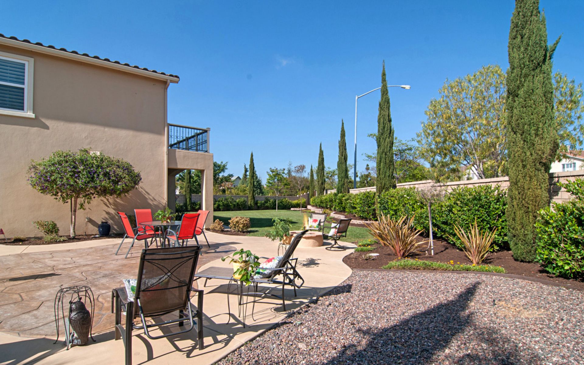 Backyard patio with red dining chairs, lounge seating, lawn, and tall cypress trees.