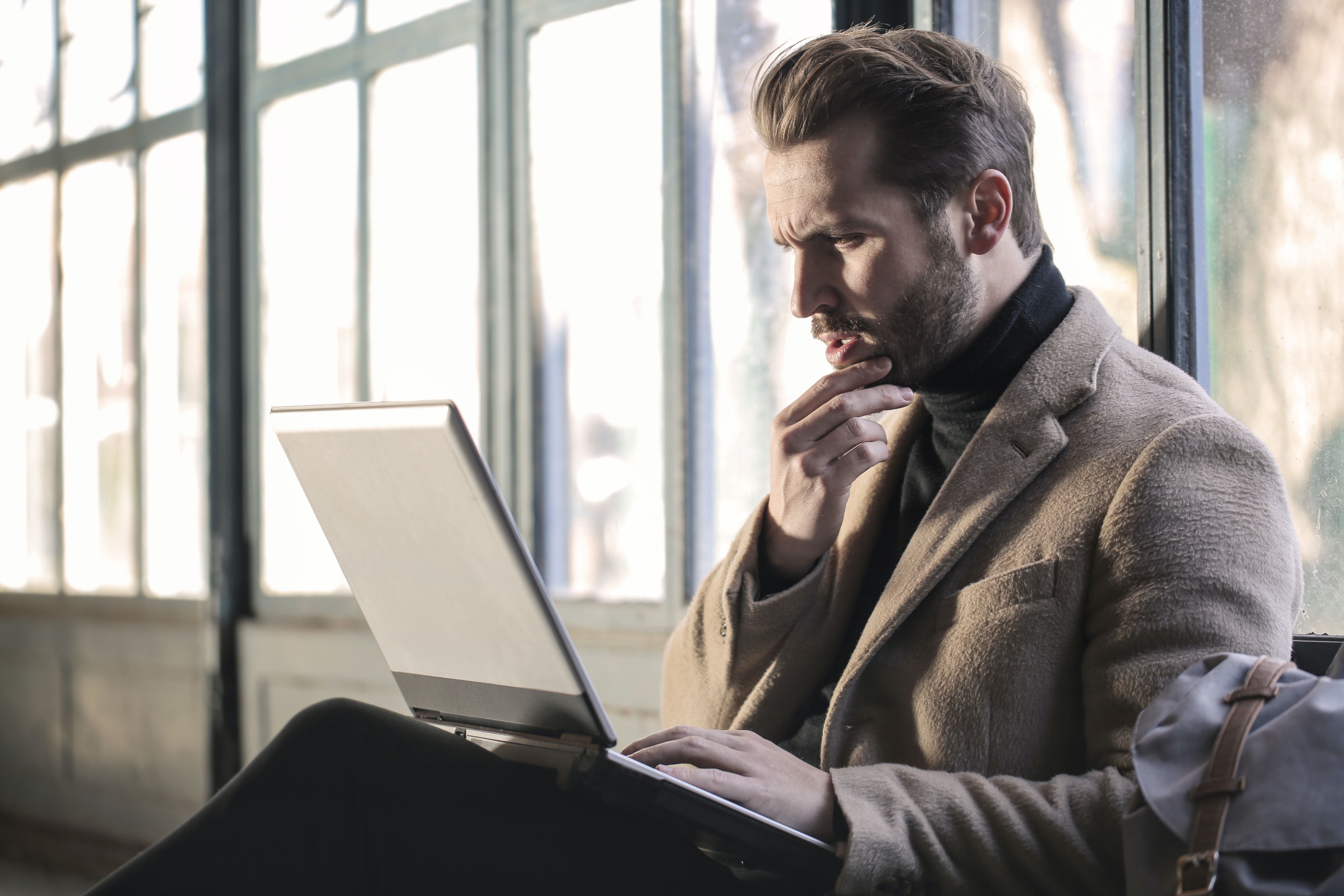 Thoughtful man studies a laptop beside large windows, resting his hand at his chin.