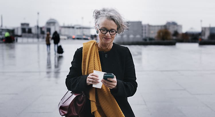 A woman wearing glasses and a yellow scarf, holding a cup and a phone. She is standing outdoors, likely in a city environment. The focus is on her casual, thoughtful demeanor.
