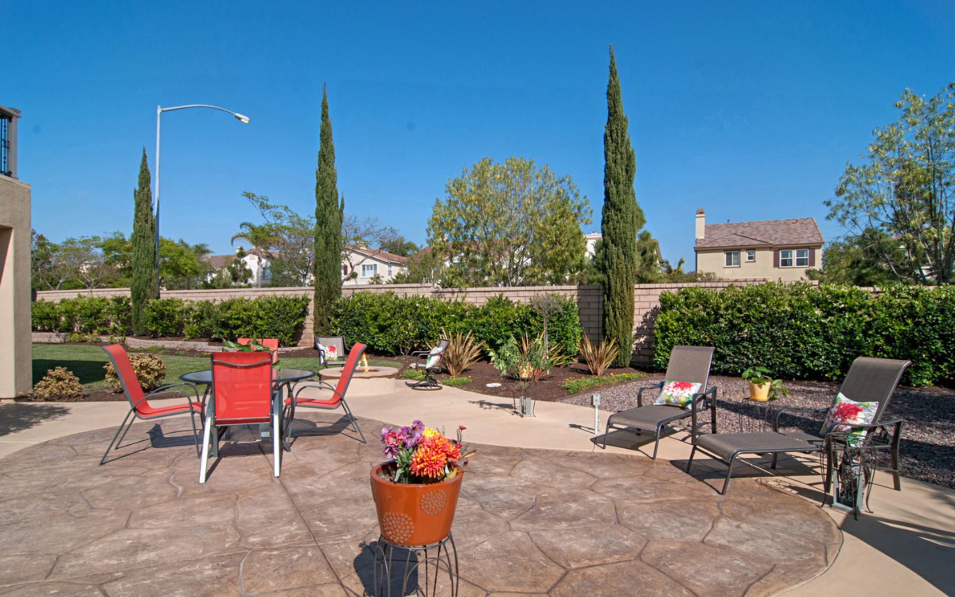 Backyard patio with stamped-concrete paving, colorful seating, potted flowers, and tall cypress trees.