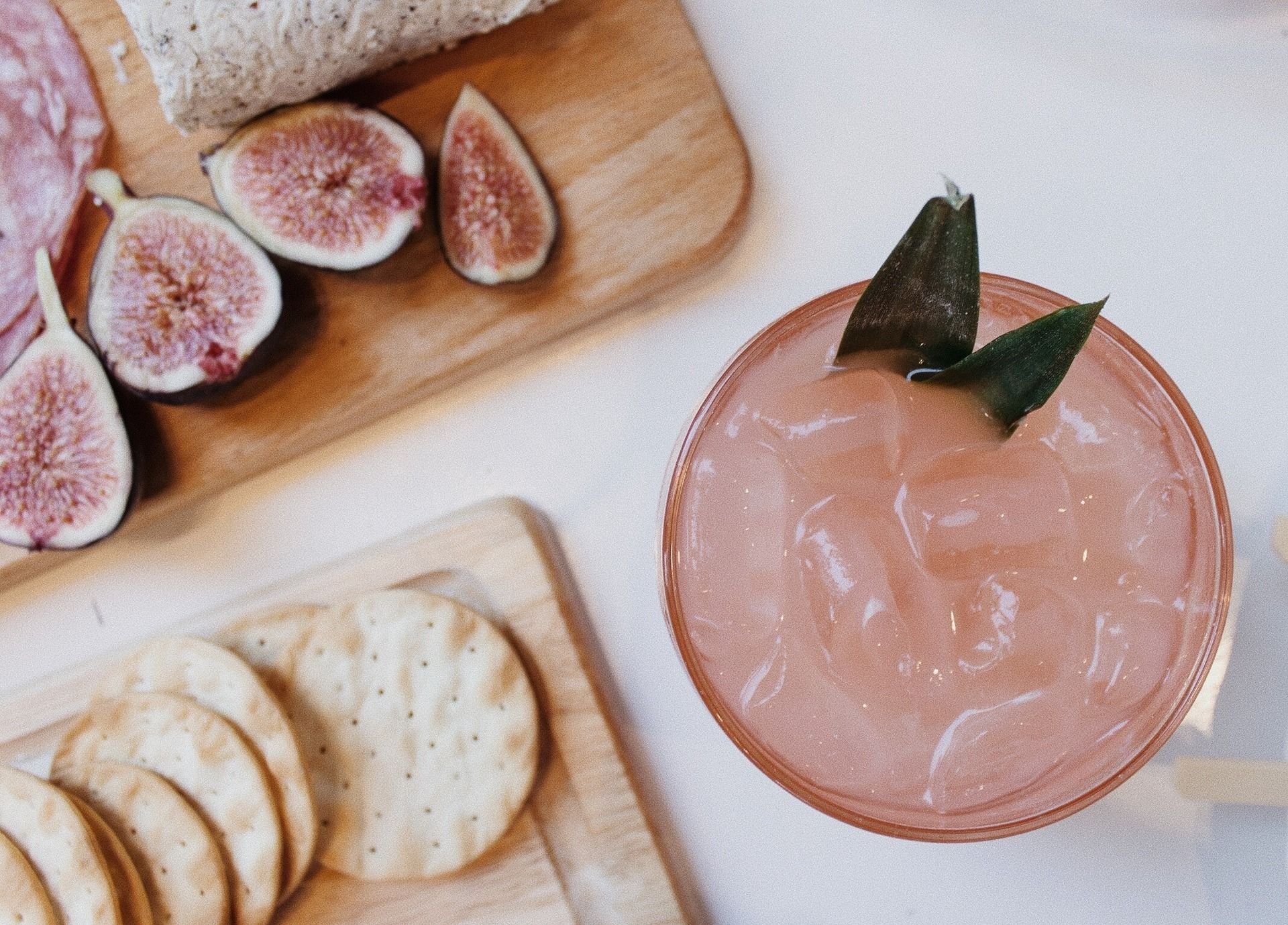 Pink iced drink with green garnish beside sliced figs, crackers, and cheese.