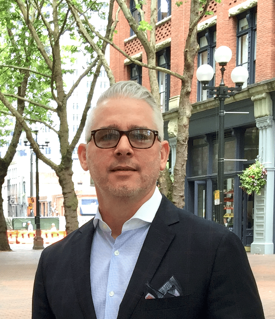 grey-haired man wearing glasses, a blue dress shirt and navy blazer standing in front of historical buildings 