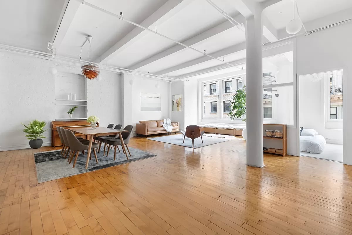 Open-plan loft living and dining area with exposed beams, hardwood floors, and a bedroom beyond.