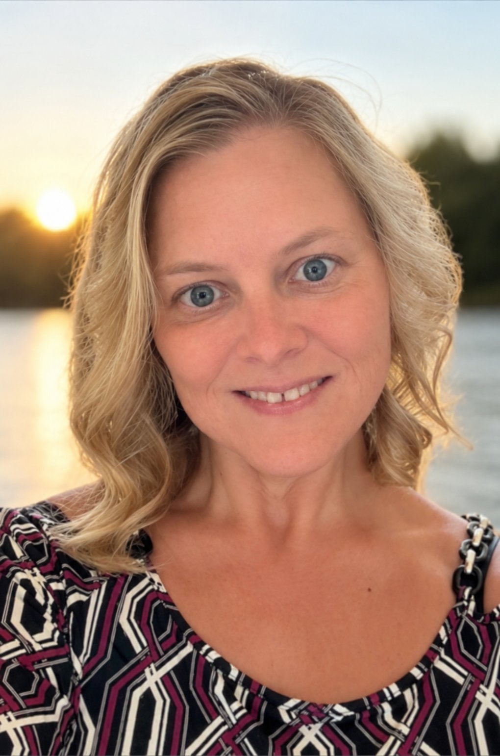Kristina Sanders smiling beside the water at sunset, wearing a black, white, and magenta patterned top.