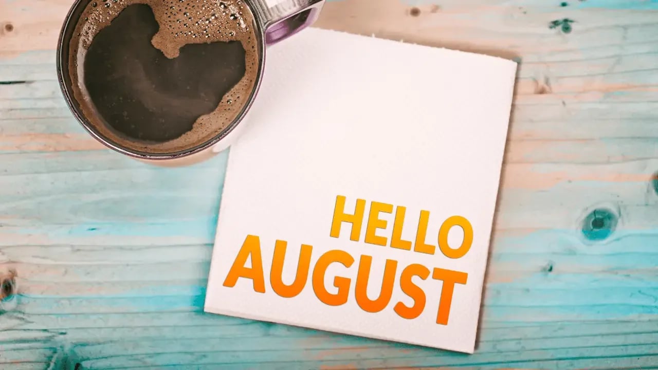 A close-up of a coffee cup on a wooden table next to a piece of paper with "HELLO AUGUST" written in orange letters. The background is a light blue wooden surface.