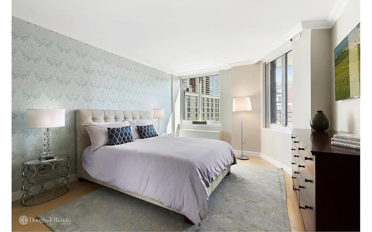 Staged bedroom with a large bed, pale blue patterned wallpaper, and tall windows overlooking nearby buildings.