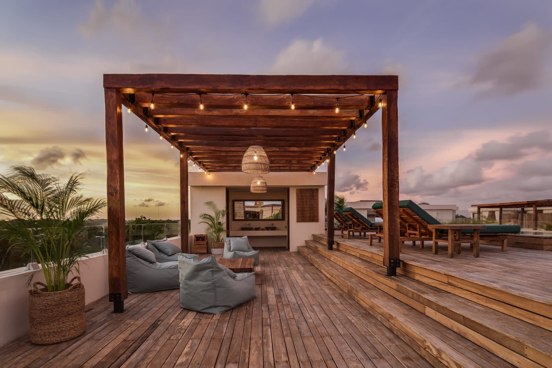 Rooftop terrace with a wooden pergola, string lights, cushioned seating, and loungers at dusk.