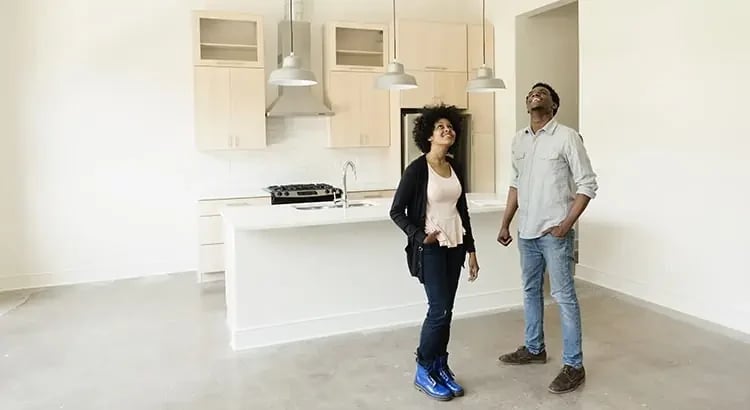 A couple standing in a modern kitchen. The kitchen has light-colored cabinets and countertops, and the couple appears to be discussing or touring the space.